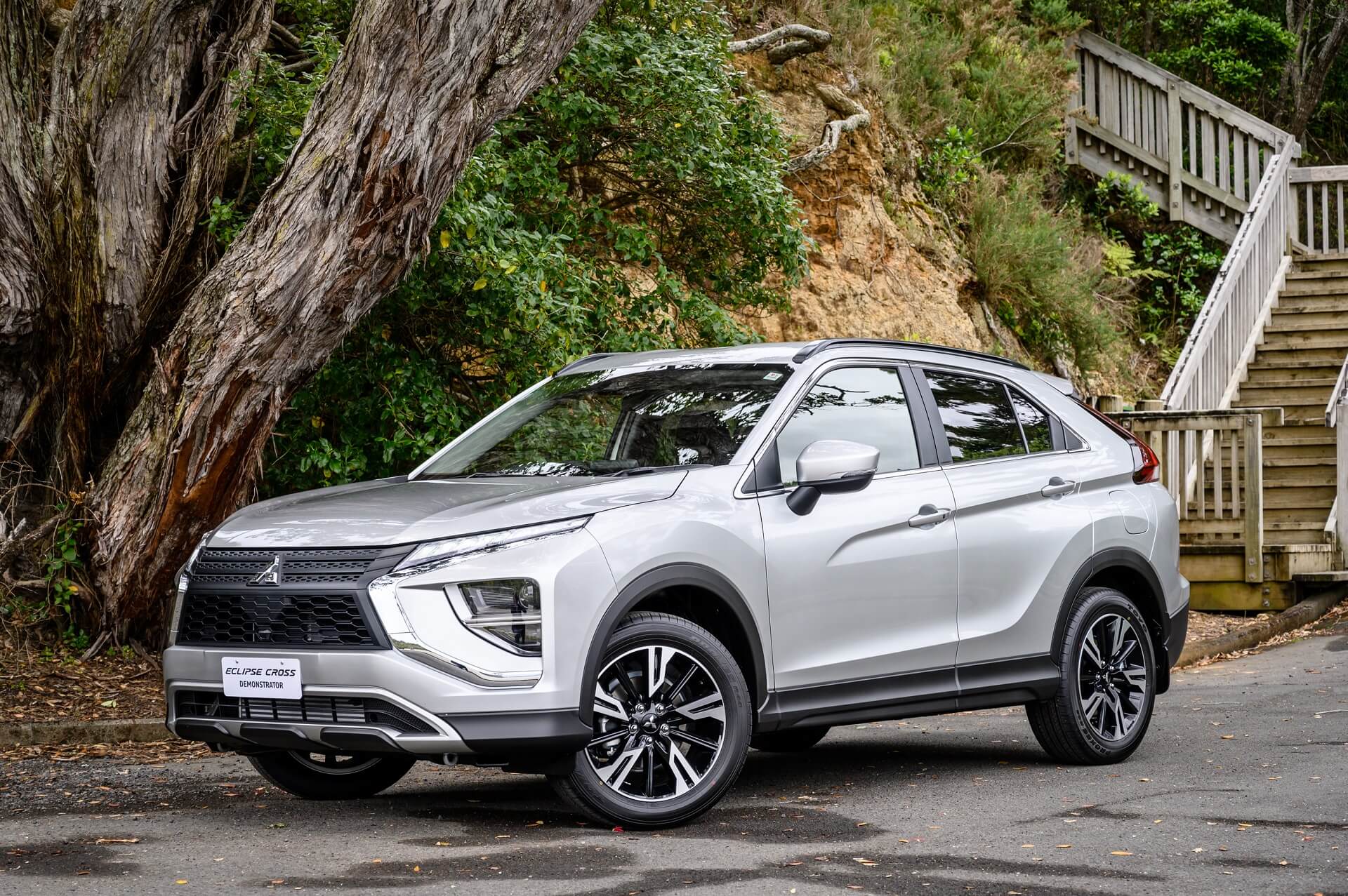 An image of a silver Mitsubishi Eclipse Cross SUV parked near a tree and some wooden stairs. 