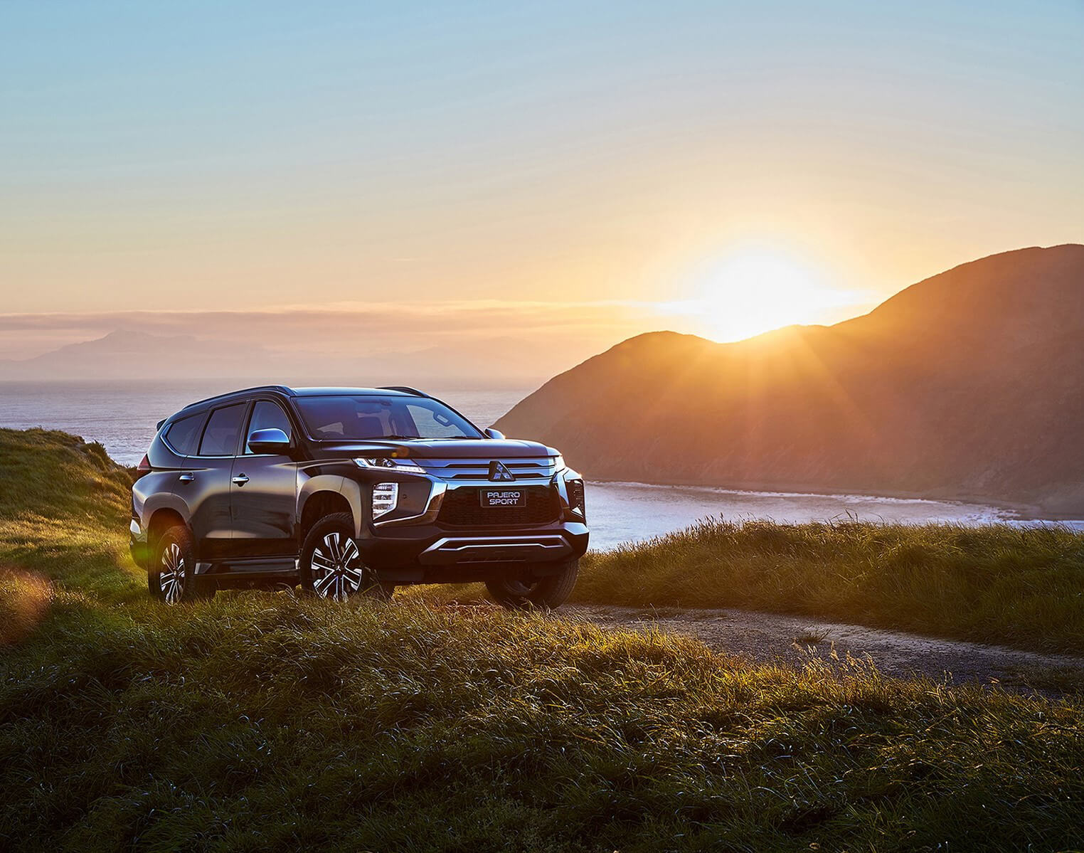A black Pajero Sport parked on a hill with sun rising from a mountain beside sea