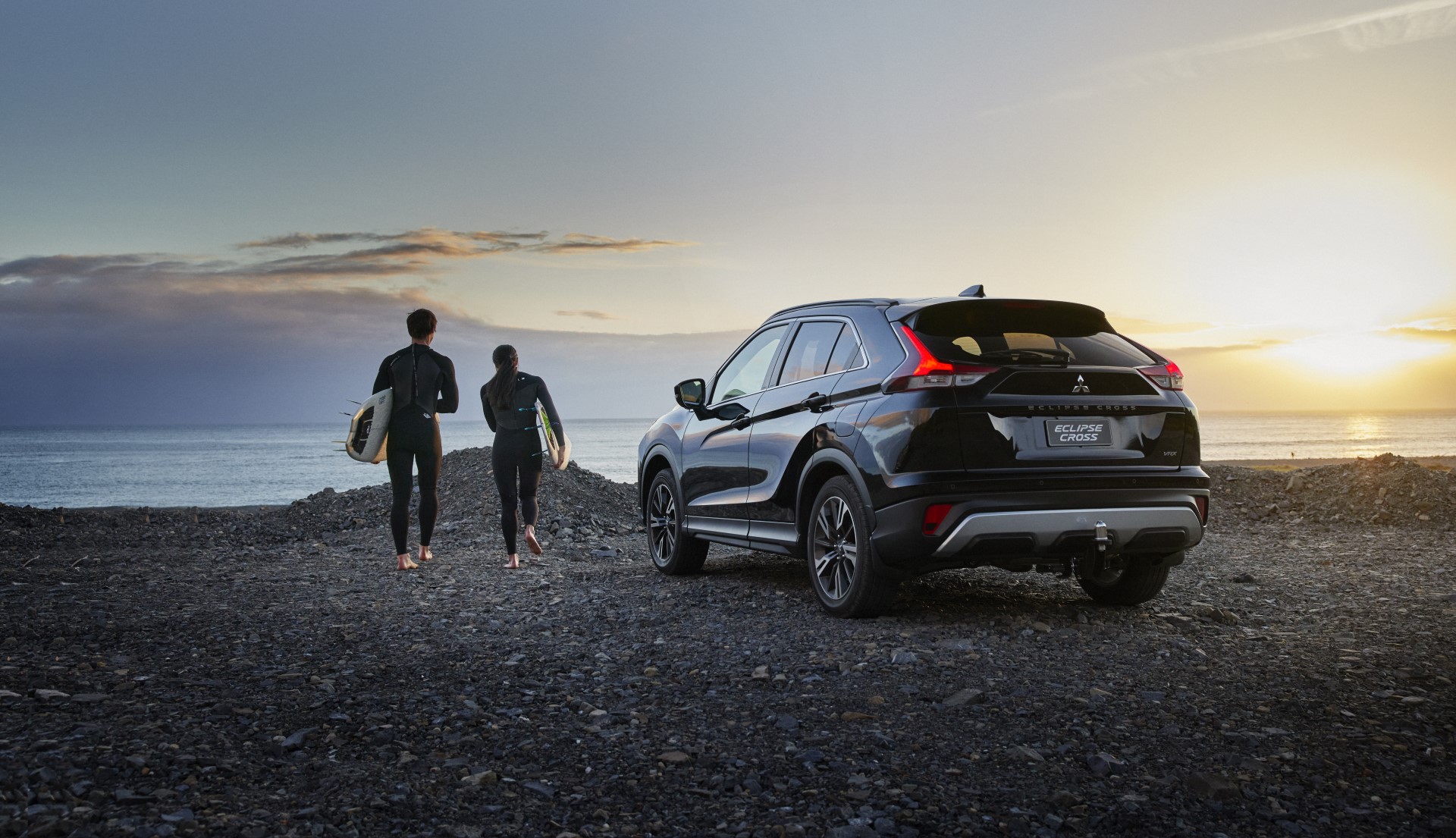 A Mitsubishi Eclipse Cross parked on a beach at sunrise with two people with wetsuits on carrying surfboards walking towards the ocean